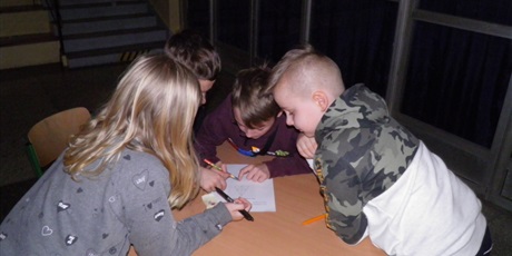 Powiększ grafikę: Czworo dzieci, 3 chłopców i dziewczynka. Siedzą przy okrągłym stoliku na którym leży kartka. Dzieci zajęte rozwiązywaniem zadań, w rękach trzymają długopisy.