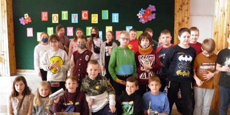 Powiększ grafikę: Klasa dzieci ubranych w różnokolorowe ubrania, patrzą w stronę obiektywu na tle dużej tablicy koloru zielonego z napisem Potyczki matematyczne.
