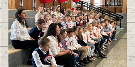 Powiększ grafikę: Grupa dzieci wraz z wychowawcami w galowych strojach  z biało-czerwonymi kotylionami przypiętymi do bluzek. Wszyscy siedzą na holu szkoły i wyglądają na zasłuchanych.