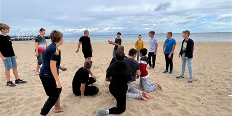 Powiększ grafikę: Grupa uczniów grających w grę "Kolanko" na plaży. W tle widać morze.