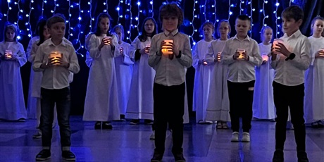 Powiększ grafikę: Dzieci trzymające w rękach świecące lampiony. Za nimi dziewczynki z lampionami w białych albach na tle kurtryny ze świateł.