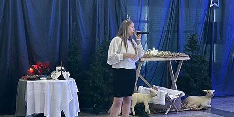 Powiększ grafikę: Dziewczyna śpiewająca kolędę a w tle stajenka, owieczki i stół ze stroikiem świątecznym.